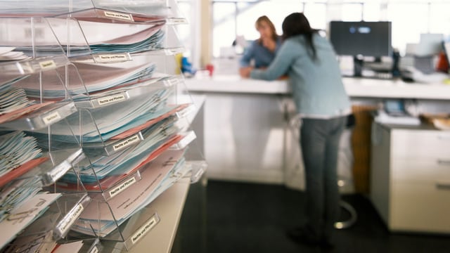 Im Vordergrund scharf Papierstapel, im Hintergrund unscharf zwei Frauen an einem Schalter im Gespräch. Im Vordergrund scharf Papierstapel, im Hintergrund unscharf zwei Frauen an einem Schalter im Gespräch.