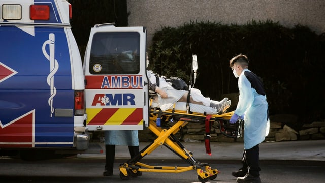 Ein Patient wird in eine Ambulanz geladen, die Nothelfer tragen Gesichtsschutz und Schutzkleidung. Ein Patient wird in eine Ambulanz geladen, die Nothelfer tragen Gesichtsschutz und Schutzkleidung.