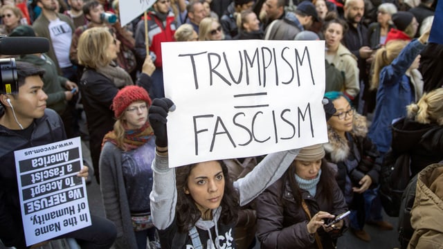 Eine Menschenmenge, aus der eine Frau hervorsticht, die ein Plakat in die Höhe streckt, auf dem steht: Trumpism = Fascism.