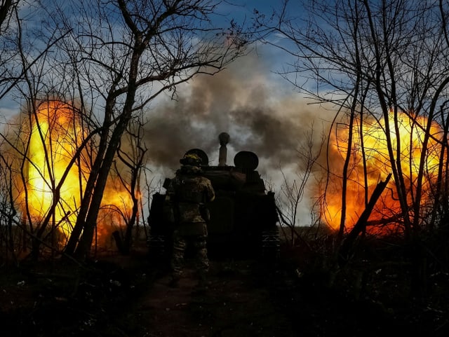 Soldaten feuern eine Panzerhaubitze auf russische Stellungen. Dabei lodern links und rechts des Geschosses flammen.