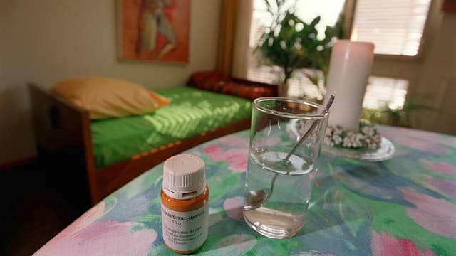 Löffel, Glas und Pentobarbital-Natrium auf Tisch stehend. Im Hintergrund ist ein Bett erkennbar Löffel, Glas und Pentobarbital-Natrium auf Tisch stehend. Im Hintergrund ist ein Bett erkennbar