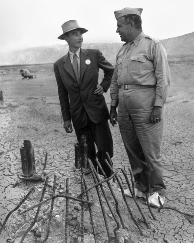 Oppenheimer im Anzug und mit Hut neben Groves in Uniform in einer Wüstenlandschaft Oppenheimer im Anzug und mit Hut neben Groves in Uniform in einer Wüstenlandschaft