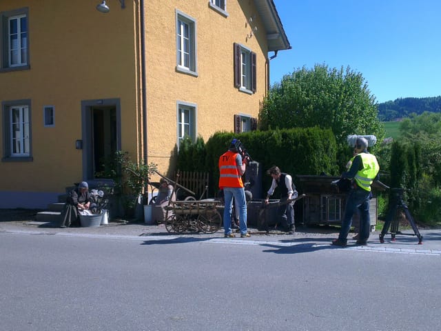 Jacqueline Büchi am waschen, Bruno Büchi am Holz hacken. Davor Kamerateam in neonfabrigen Westen.