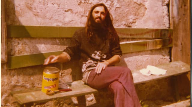 Ein Mann mit langen Haaren und Vollbart sitzt auf einer Bank. Ein Mann mit langen Haaren und Vollbart sitzt auf einer Bank.