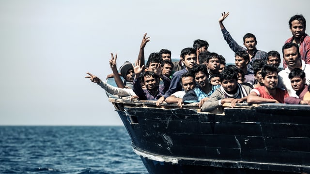 Menschen dichtegedrängt auf einem Holzkahn auf offenem Meer, manche winken mit den Händen. Menschen dichtegedrängt auf einem Holzkahn auf offenem Meer, manche winken mit den Händen.