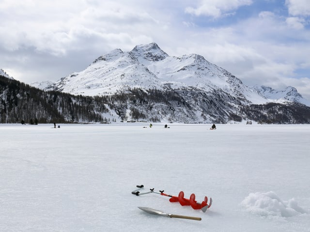 Gefrorener See mit Eisfischern. Im Hintergrund ein verschneiter Berg und nur wenige Wolkenlücken.