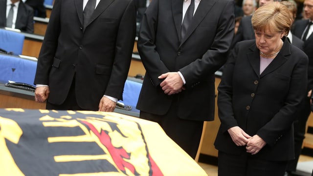 Merkel vor einem mit der deutschen Flagge geschmückten Sarg. Merkel vor einem mit der deutschen Flagge geschmückten Sarg.