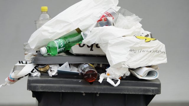 Eine schwarze überfülte Abfalltonne. Plastiksäcke und PET-Flaschen fallen heraus. Eine schwarze überfülte Abfalltonne. Plastiksäcke und PET-Flaschen fallen heraus.