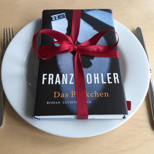 Das Buch «Das Päckchen» liegt auf einem weissen Teller mit einer Geschenkmasche drum Das Buch «Das Päckchen» liegt auf einem weissen Teller mit einer Geschenkmasche drum