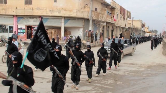 Schwarz gekleidete und bewaffnete Männer marschieren in zwei Reihen durch eine Ortschaft. Schwarz gekleidete und bewaffnete Männer marschieren in zwei Reihen durch eine Ortschaft.