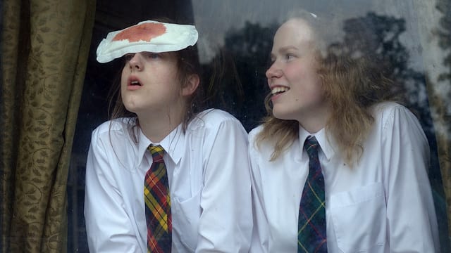 Zwei Mädchen in Schuluniform stehen an einem Fenster, einer klebt eine blutige Binde auf der Stirn. Zwei Mädchen in Schuluniform stehen an einem Fenster, einer klebt eine blutige Binde auf der Stirn.