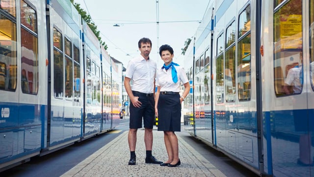 Eine Trampilotin und ein Trampilot in blauem Jupe und kurzen Hosen, mit weisser Blue und Hemd, zwischen zwei Tram. Eine Trampilotin und ein Trampilot in blauem Jupe und kurzen Hosen, mit weisser Blue und Hemd, zwischen zwei Tram.