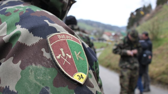 Mann mit Armeeabzeichen steht im Vordergrund. Im Hintergrund Armeeangehörige am Arbeiten. Mann mit Armeeabzeichen steht im Vordergrund. Im Hintergrund Armeeangehörige am Arbeiten.