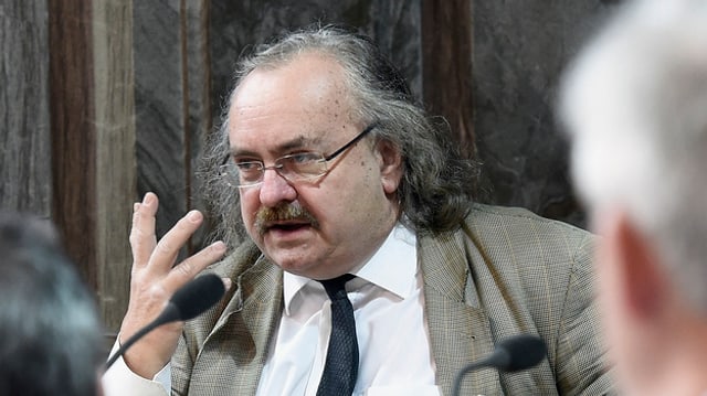 Älterer Mann mit Brille hebt die Hand vor einem Mikrofon. Älterer Mann mit Brille hebt die Hand vor einem Mikrofon.