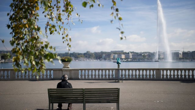 Ein Mann mit Kopfhörern sitzt auf einer Bank an der Promenade am Genfersee, rechts der Jet d'eau.