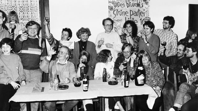 Eine Gruppe von fröhlichen Menschen sitzen dund stehend an einem Tisch, hinten ein Grünen-Transparent Eine Gruppe von fröhlichen Menschen sitzen dund stehend an einem Tisch, hinten ein Grünen-Transparent