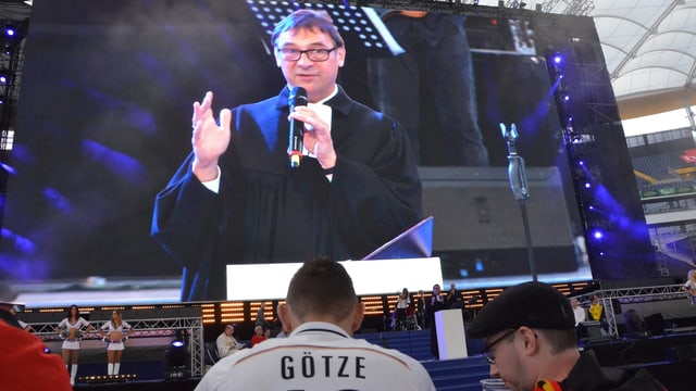 Messe im Stadion vor dem Spiel, Fans stehen vor der Riesenleinwand, darauf predigt ein Pfarrer. Messe im Stadion vor dem Spiel, Fans stehen vor der Riesenleinwand, darauf predigt ein Pfarrer.