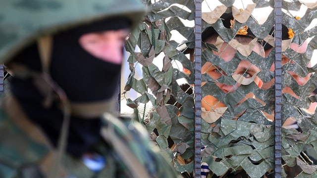 Ein Vermummter steht vor einem Gitter, dahinter blicken zwei Uniformierte nach draussen. Ein Vermummter steht vor einem Gitter, dahinter blicken zwei Uniformierte nach draussen.