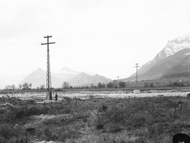 Eine historische schwarz-weiss Foto mit einem offen Feld und einem Strommasten, dahinter die Berge. 