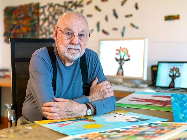 Ein älterer Herr mit Glatze und blauem Pullover sitzt an einem Schreibtisch, auf dem bunte Bilder liegen.