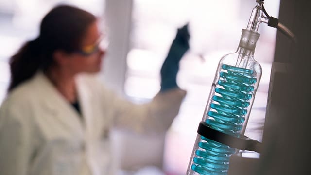 Labor: Flasche mit blauer Flüssigkeit, im Hintergrund eine Frau mit Schutzbrille 
