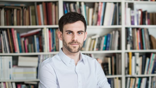 Bild von einem Mann vor einem Büchergestell. Bild von einem Mann vor einem Büchergestell.