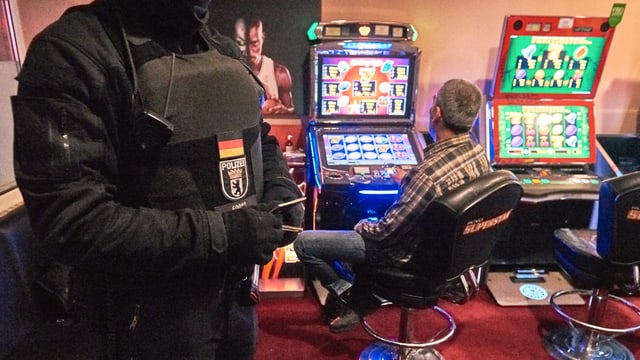 Mann sitzt vor Spielautomat. Daneben steht ein Polizist in Schutzkleidung. Mann sitzt vor Spielautomat. Daneben steht ein Polizist in Schutzkleidung.
