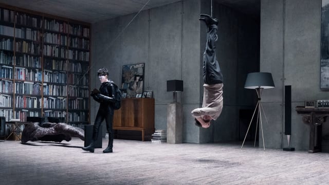 In einem Haus, die Einrichtung ist teuer, aber trist. An der linken Wand ein riesiges Bücherregal. Davor liegt die moderne Steinstatue eines Engels umgekippt am Boden. Von der Statue führt ein Seil über einen Haken an der Deckezu einem Mann. Das Seil ist um seine Füsse gewickelt, er hängt kopfüber in der Luft. Vor ihm steh eine Frau mit kurzen Haaren. In einem Haus, die Einrichtung ist teuer, aber trist. An der linken Wand ein riesiges Bücherregal. Davor liegt die moderne Steinstatue eines Engels umgekippt am Boden. Von der Statue führt ein Seil über einen Haken an der Deckezu einem Mann. Das Seil ist um seine Füsse gewickelt, er hängt kopfüber in der Luft. Vor ihm steh eine Frau mit kurzen Haaren.