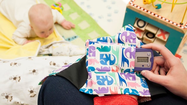 Ein Baby liegt auf dem Boden, davor hält die Mutter ein kleines Gerät in der Hand. Ein Baby liegt auf dem Boden, davor hält die Mutter ein kleines Gerät in der Hand.