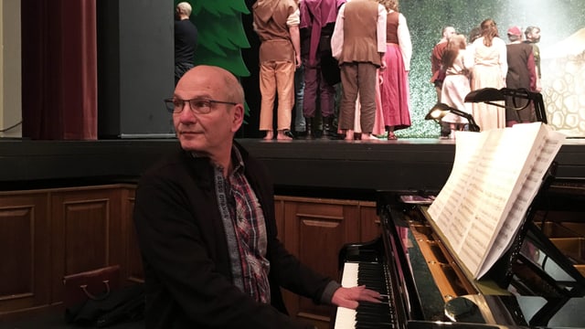 Ein Mann sitzt am Klavier und spielt, im Hintergrund stehen Kinder auf einer Theaterbühne. Ein Mann sitzt am Klavier und spielt, im Hintergrund stehen Kinder auf einer Theaterbühne.