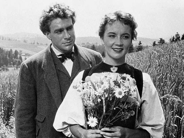 Schwarz-Weiss-Aufnahme: Liselotte Pulver in Bernertracht und mit Blumenstrauss. Dahinter steht Hannes Schmidhauser. Schwarz-Weiss-Aufnahme: Liselotte Pulver in Bernertracht und mit Blumenstrauss. Dahinter steht Hannes Schmidhauser.