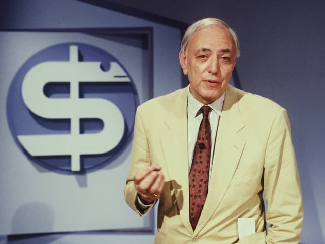 Seriöser, älterer Fernsehmoderator vor einem stilisierten Aeskulap-Stab Seriöser, älterer Fernsehmoderator vor einem stilisierten Aeskulap-Stab