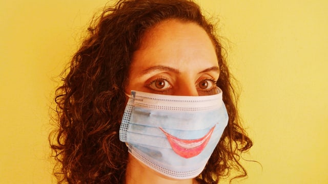 Eine Frau trägt einen Mundschutz. Auf diesem ist ein lachender Mund gezeichnet. Eine Frau trägt einen Mundschutz. Auf diesem ist ein lachender Mund gezeichnet.