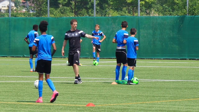 Ein Trainer in schwarzen kurzen Hosen gibt Anweisungen an Fussballjunioren, die auf dem Platz herumstehen mit einem Ball am Fuss. Ein Trainer in schwarzen kurzen Hosen gibt Anweisungen an Fussballjunioren, die auf dem Platz herumstehen mit einem Ball am Fuss.