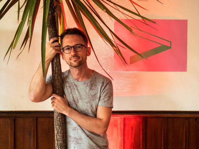 Mann mit Brille lehnt lächelnd an einer Palme in einem Raum Mann mit Brille lehnt lächelnd an einer Palme in einem Raum