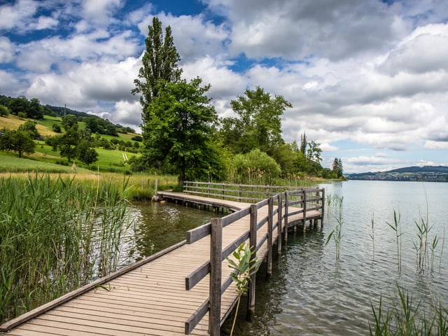 Ein Holzsteg führt am Ufer des Sees entlang, auf seiner glatten Wasseroberfläche spiegeln sich Wolken.