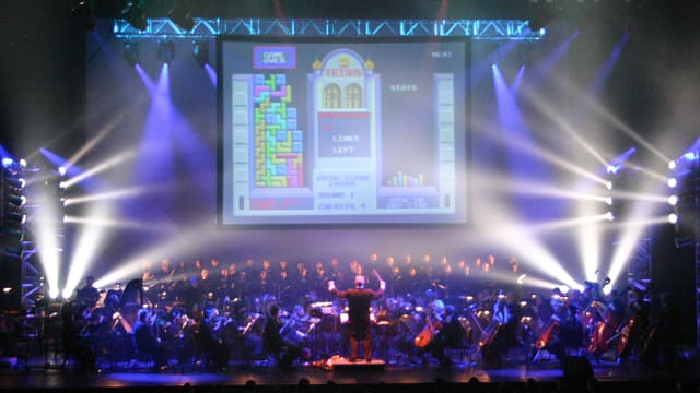 Auf einer Bühne spielt ein Orchester, im Hintergrund eine Leinwand auf der das Spiel "Tetris" zu sehen, Auf einer Bühne spielt ein Orchester, im Hintergrund eine Leinwand auf der das Spiel "Tetris" zu sehen,