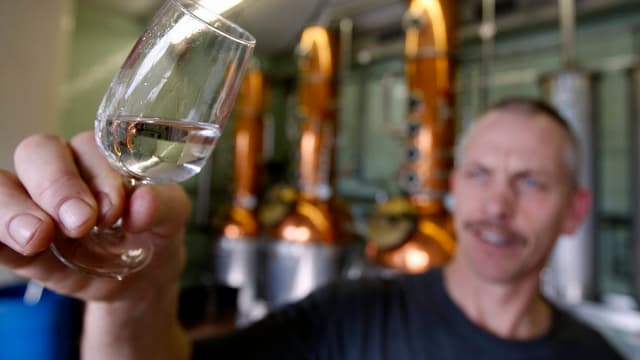 Ein Mann hält in einer Kleinbrennerei ein Gläschen Schnaps in die Kamera, im Hintergrund die Brennhäfen. Ein Mann hält in einer Kleinbrennerei ein Gläschen Schnaps in die Kamera, im Hintergrund die Brennhäfen.