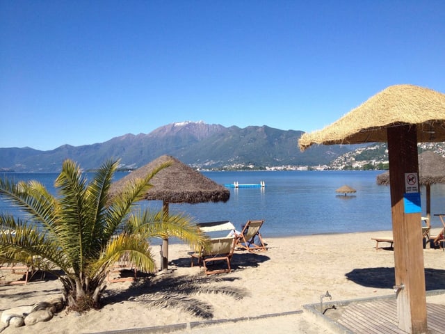 Palme auf Sandstrand, Liegestühle und Sonnenschirme aus Stroh. Ein See und Berge bei schönstem Sommerwetter. Palme auf Sandstrand, Liegestühle und Sonnenschirme aus Stroh. Ein See und Berge bei schönstem Sommerwetter.