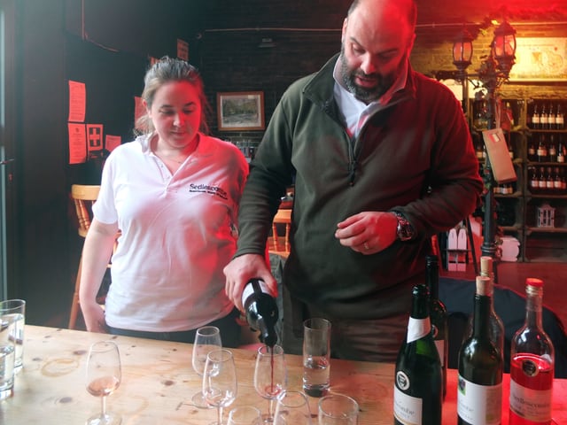 Ein frau und ein Mann stehen an einem Tisch. Er schenkt ein Glas Rotwein ein, auf dem Tisch stehen drei weitere Weinflaschen Ein frau und ein Mann stehen an einem Tisch. Er schenkt ein Glas Rotwein ein, auf dem Tisch stehen drei weitere Weinflaschen