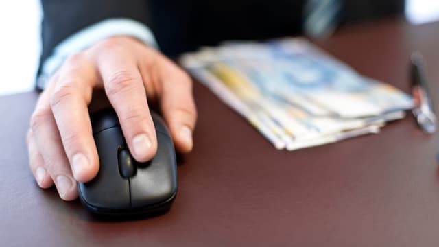Ein Mann mit einer Computermaus in der Hand sitzt an einem Tisch, auf dem einige Hunderternoten liegen. Ein Mann mit einer Computermaus in der Hand sitzt an einem Tisch, auf dem einige Hunderternoten liegen.