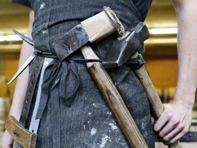 Bildausschnitt: Am Gürtel einer Arbeitsschürze hängen schwere Hämmer und Metallwerkzeuge.