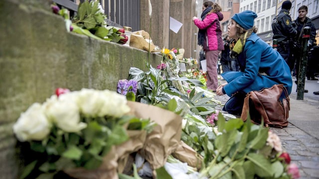 Blumen liegen vor einem Gittertor, Menschen kauern daneben, im Hintergrund bewaffnete Polizisten.