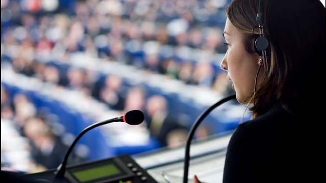 Eine Übersetzerin mit Kopfhörer spricht in ein Mikrofon während einer Debatte. Eine Übersetzerin mit Kopfhörer spricht in ein Mikrofon während einer Debatte.