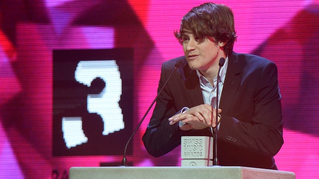 Ein junger Mann mit Anzug und glattem Haar, das über Stirn und Augen fällt, steht an einem Pult mit zwei Mikrofonen. Vor ihm steht ein kleiner Betonklotz mit der Inschrift Swiss Music Awards. Im Hintergrund ist das Zeichen von Radio SRF 3 zu sehen. Ein junger Mann mit Anzug und glattem Haar, das über Stirn und Augen fällt, steht an einem Pult mit zwei Mikrofonen. Vor ihm steht ein kleiner Betonklotz mit der Inschrift Swiss Music Awards. Im Hintergrund ist das Zeichen von Radio SRF 3 zu sehen.