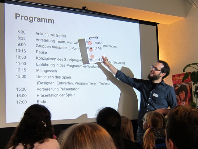 Ein Mann zeigt mit einem Notizblock ein vom Hellraumprojektor auf eine Leinwand projiziertes Tagesprogramm.