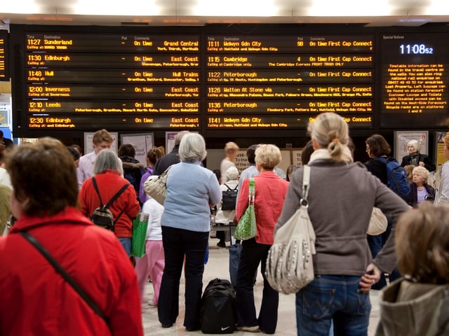 Viele Menschen stehen mit Blick auf eine orange leuchtende, elektronische Anzeigetafel, in einer Bahnhofshalle und warten.. Viele Menschen stehen mit Blick auf eine orange leuchtende, elektronische Anzeigetafel, in einer Bahnhofshalle und warten..