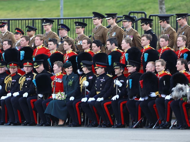 Herzogin Kate und PRinz William sitzen umgeben von Männern in Uniform auf einer Bank.