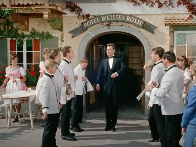 Kellner tritt aus Eingang zum weissen Rössl heraus und wird von Hilfskellnern flankiert. Kellner tritt aus Eingang zum weissen Rössl heraus und wird von Hilfskellnern flankiert.