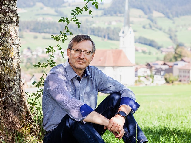 Urs Corradini sitzt neben einem Baum. Im Hintergrund ist eine Kirche zu sehen. Urs Corradini sitzt neben einem Baum. Im Hintergrund ist eine Kirche zu sehen.